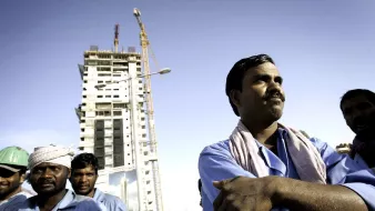 L'image montre un groupe d'ouvriers sur un chantier de construction. Au premier plan, un homme est au centre de l'image, les bras croisés et affichant une expression confiante. À l'arrière-plan, on voit un bâtiment en construction, avec des grues qui travaillent. Les ouvriers portent des vêtements de travail, et certains d'entre eux portent des casques de sécurité. Le ciel est dégagé, suggérant une journée ensoleillée. L'ensemble de la scène évoque l'atmosphère d'un site de construction animé avec des travailleurs engagés dans leur tâches.