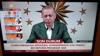 L'image montre un homme en train de prendre la parole lors d'une conférence, probablement un discours politique. Il porte une veste verte et se tient devant un grand fond décoré, avec ce qui semble être un symbole national ou institutionnel. En bas de l'écran, il y a un bandeau rouge qui indique "SON DURUM" et mentionne "CUMHURBAŞKANI ERDOĞAN" avec un message sur des réformes importantes. Sur le côté gauche, des informations sur des élections et des résultats sont affichées, suggérant que l'événement est lié à des élections en Turquie.