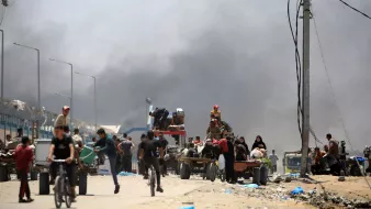 L'image montre une scène chaotique dans un environnement urbain. On peut voir des gens marchant et roulant à vélo le long d'une route, tandis qu'un groupe semble être occupé à charger ou décharger des marchandises sur des chariots. En arrière-plan, de la fumée s'élève, suggérant qu'un incendie ou une explosion a eu lieu. L'ambiance semble tendue et désordonnée, avec des câbles électriques visibles et des structures urbaines en arrière-plan.