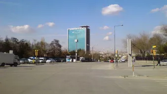 L'image montre une vue d'une rue avec un bâtiment moderne en arrière-plan. Ce bâtiment, de grande hauteur, a une façade en verre qui reflète le ciel. Le ciel est clair avec quelques nuages dispersés. On peut voir des voitures garées et circulant sur la route, ainsi que des arbres sur le côté. L'ensemble donne une impression de ville moderne et animée.