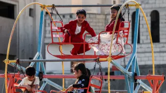 L'immagine mostra un'area giochi all'aperto, con una giostra colorata. Alcuni bambini sono seduti su seggiolini rossi mentre si divertono. Uno dei bambini indossa un completo rosso e sembra divertirsi molto, mentre un'altra bambina, vestita con un abito bianco con fiori, osserva da un'altezza superiore. Gli altri bambini sono anche loro intenti a giocare, creando un'atmosfera di gioia e spensieratezza in un contesto urbano.