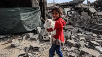 Sur cette image, on voit une jeune fille souriante portant un chat blanc dans ses bras. Elle est habillée d'un manteau rouge et d'un jean bleu. La scène se déroule au milieu de décombres, avec des bâtiments détruits et des débris tout autour. Le ciel est partiellement visible et semble être couvert de nuages. L'image donne une impression de résilience et d'espoir malgré un environnement de destruction.