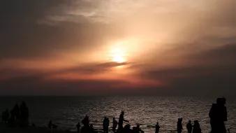 L'image montre une plage au crépuscule, avec des nuages qui tamisent la lumière du soleil couchant. De nombreuses silhouettes de personnes se déplacent le long du rivage, profitant de la scène. La mer reflète les couleurs douces du ciel, créant une atmosphère tranquille et paisible. Les silhouettes des individus ajoutent une dynamique à l'image, suggérant une ambiance conviviale et joyeuse.