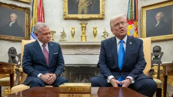 L'image montre deux hommes assis dans une pièce élégante, probablement dans un bureau officiel. L'un d'eux, portant un costume sombre, regarde avec une expression sérieuse, tandis que l'autre, en costume clair avec une cravate bleue, parle. Des drapeaux et des tableaux anciens décorent la pièce. Une table en bois est visible devant eux.