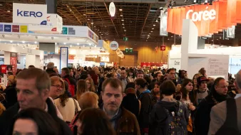 L'image montre une foule dense de personnes rassemblées dans un espace d'exposition, probablement lors d'un événement ou d'une foire. On peut voir des stands avec des bannières et des affiches en arrière-plan, créant une atmosphère dynamique. Les gens sont variés en termes de styles vestimentaires et d'âges, indiquant un mélange diversifié de visiteurs. L'éclairage est lumineux, mettant l'accent sur l'activité animée qui se déroule dans ce lieu.