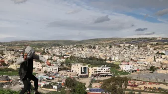 Nell'immagine si vede un giovane che si sta arrampicando su una roccia, con una città sullo sfondo. La città è composta da edifici di diversi piani, molti dei quali di colore chiaro. L'area è circondata da colline verdi e il cielo è parzialmente nuvoloso, suggerendo un clima variabile. Si possono notare anche alcune strutture agricole e aree di terreno aperto in primo piano.
