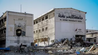 The image depicts a scene of devastation, likely in a war-torn area. In the foreground, there is rubble and debris from damaged buildings, suggesting destruction from conflict. A partially destroyed building is visible, with the name "Al Azhar University - Gaza" written on it in Arabic, indicating it is an educational institution. In the foreground, two individuals on bicycles navigate through the debris, highlighting the struggle of daily life in such an environment. The overall atmosphere conveys a sense of loss and destruction, with remnants of architecture marked by chaos.