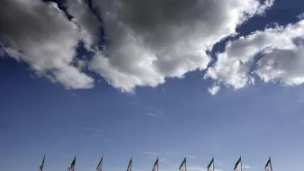 The image features a clear blue sky with fluffy white clouds. In the foreground, several flags, likely representing Mexico, are lined up. The flags prominently display the colors of the Mexican flag, which includes green, white, and red, along with the national emblem. The overall scene conveys a sense of patriotism against a serene backdrop.