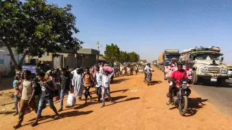 L'image montre une scène de rue animée dans une zone urbaine. Sur le côté gauche, on voit un arbre et des bâtiments, tandis que des gens marchent le long de la route en terre. Parmi eux, certains portent des sacs ou des charges sur la tête. Des véhicules, y compris des camions et des motos, circulent sur la route, créant une ambiance dynamique. L'éclairage semble être en plein jour, suggérant une activité quotidienne typique dans cette communauté.