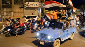 در این تصویر، گروهی از جوانان در یک خیابان در حال شادی و جشن هستند. آنها بر روی یک خودروِ آبی رنگ نشسته‌اند و پرچم عراق را در دست دارند. تعدادی موتورسیکلت نیز در کنار خودرو در حال حرکت هستند. جوانان با شور و هیجان به جشن و شادی پرداخته و فضای پرانرژی و شادابی را ایجاد کرده‌اند. در پس‌زمینه، ساختمان‌ها و فروشگاه‌هایی دیده می‌شوند که به شلوغی و نشاط این صحنه افزوده‌اند.