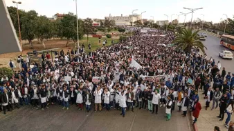 L'image montre une grande foule de manifestants rassemblés dans une rue, probablement pour une manifestation ou une protestation. Beaucoup de participants portent des blouses blanches, ce qui pourrait indiquer qu'ils sont des professionnels de la santé ou des étudiants en médecine. Ils agitent des panneaux et des drapeaux, exprimant leurs revendications. L'environnement semble urbain, avec des arbres et des bâtiments en arrière-plan. L'ambiance générale paraît mobilisée et solidaire.