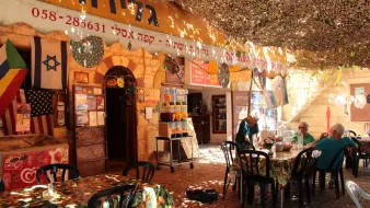 L'image montre un café ou une taverne en plein air, avec des tables et des chaises noires disposées sur un sol en pierre. Le lieu semble très accueillant, avec des nappes colorées sur les tables. Au-dessus, un toit en toile de camouflage laisse passer un peu de lumière. On peut voir des drapeaux accrochés aux murs, probablement des symboles de différentes nationalités. Des personnes assises discutent tranquillement dans cet environnement convivial. L'architecture est typique des bâtiments anciens, ajoutant une touche authentique à l'ambiance.