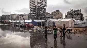 Un campo profughi con tende, bambini giocano e un edificio in rovina sullo sfondo.