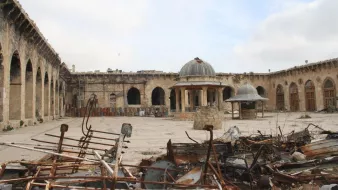 L'image montre un site historique en ruines. On peut voir une cour spacieuse entourée de murs en pierre, avec des arches et des colonnes qui témoignent d'une architecture ancienne. Au centre, il y a deux structures en forme de dômes. Le sol est recouvert de débris métalliques et de morceaux de ferraille, suggérant que le lieu a été touché par des événements destructeurs. Le ciel est nuageux, ajoutant à l'atmosphère de désolation.