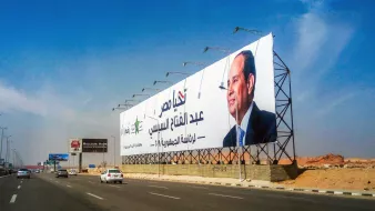 The image depicts a large billboard along a highway featuring a prominent figure, likely a politician, with text in Arabic. The background shows a clear blue sky, and the area appears to be urban, with vehicles on the road. The billboard is designed to attract attention, showcasing both the individual's image and a message or campaign slogan.