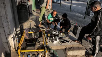 Cette image montre un groupe de personnes, principalement des hommes, rassemblées autour d'un générateur et de plusieurs chargeurs de téléphone portable. Ils se trouvent à l'extérieur, probablement sur un trottoir ou une zone piétonne pavée. Plusieurs téléphones portables sont connectés aux chargeurs qui sont branchés au générateur. À l'arrière-plan, on peut voir une voiture garée et des bâtiments. L'atmosphère semble indiquer une situation où les gens utilisent un générateur pour charger leurs appareils en l'absence d'une source d'électricité régulière.