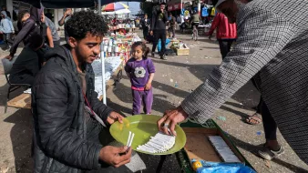 L'image montre une scène de marché dans une rue animée. Un jeune homme est assis sur un banc, tenant une assiette ronde verte sur ses genoux, où semblent être disposés des articles, probablement des bonbons ou des bâtonnets. Un homme plus âgé s'approche de lui, tenant quelque chose dans sa main, peut-être en train de lui faire une offre ou de lui donner des articles. En arrière-plan, il y a des gens qui se déplacent, ainsi que des étals avec divers produits. Une petite fille se tient à proximité, observant la scène. Les parasols créent une ambiance de marché, tandis que l'atmosphère semble vivante et animée.