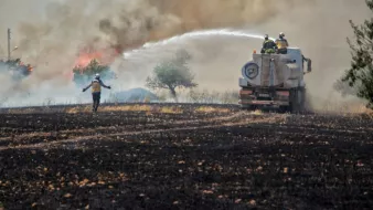 L'image montre une scène de lutte contre un incendie. On voit des pompiers en action, utilisant un camion-citerne pour éteindre les flammes qui s'élèvent dans la fumée. Au premier plan, un pompier semble se diriger vers le feu, tandis que l'arrière-plan montre un paysage noirci, témoignant des dégâts causés par l'incendie. Les arbres sont entourés de fumée, créant une atmosphère de tension et d'urgence.