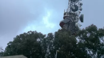 L'image montre une tour de télécommunication élevée, dotée de plusieurs antennes et équipements en haut. Elle est entourée de grands arbres verts et se dresse à côté d'un bâtiment plus petit, probablement un centre d'opérations ou de maintenance. Le ciel est nuageux, ce qui donne une ambiance légèrement grise et sombre à la scène.