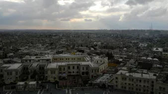 L'image montre une vue panoramique d'une ville, probablement une ancienne ville, avec des bâtiments en pierre grise et une architecture typique du Moyen-Orient. Le ciel est nuageux, avec des rayons de lumière qui percent à travers les nuages, créant une atmosphère dramatique. À l'arrière-plan, on peut apercevoir d'autres structures urbaines et des mosquées, indiquant une richesse historique. L'ambiance semble à la fois paisible et chargée de mélancolie.