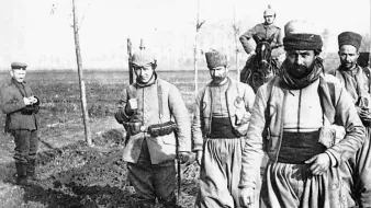 The image depicts a group of soldiers walking along a muddy path. They are dressed in military uniforms typical of the early 20th century, with notable features like baggy trousers and headgear, indicating a specific cultural or historical context. One soldier is on horseback in the background, while another man stands to the side, possibly observing. The environment appears rural, with sparse trees lining the path. The overall atmosphere conveys a sense of movement and purpose in a military setting.