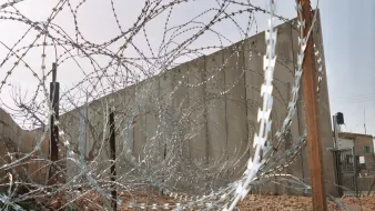 L'image montre un paysage marqué par des barbelés disposés en spirale et un mur en béton en arrière-plan. Le terrain est rocheux et dépouillé, ce qui contribue à une ambiance austère et sécurisée. Les barbelés créent une impression de barricade ou de clôture, suggérant une séparation ou un isolement. Le ciel au-dessus est clair, ajoutant un contraste avec les éléments de sécurité présents dans le paysage.