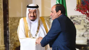 The image depicts two men in formal attire shaking hands. One man is wearing traditional Saudi clothing, including a white thobe and a black and white headscarf, adorned with a gold sash. The second man is dressed in a dark suit. They appear to be engaged in a cordial greeting, likely in a diplomatic or official context. In the background, there are hints of decor that suggest an elegant setting, possibly within a government building. One of the men is also positioned near a flag, indicating a connection to national representation.