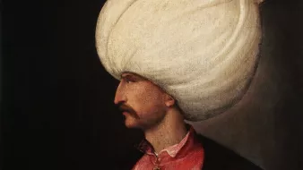 L'image représente un homme en profil, portant un turban blanc imposant. Il est habillé d'une tunique rose ornée de boutons et d'un veston noir, ce qui lui donne un aspect richement décoré. Son visage est finement dessiné, mettant en valeur sa moustache. Il tient un bâton dans une main, ce qui suggère une position de pouvoir ou d'autorité. L'arrière-plan est sombre, ce qui fait ressortir les détails de sa tenue et de son expression.