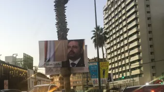 L'image montre une vue urbaine avec un grand panneau publicitaire affichant le visage d'un homme en costume, probablement un homme politique, et des arbres de palmiers sur le côté. En arrière-plan, on peut voir un immeuble de grande hauteur avec des enseignes, et d'autres panneaux d'affichage autour. La lumière du soir donne une ambiance douce à la scène. Les voitures sont visibles sur la route, suggérant une circulation urbaine.