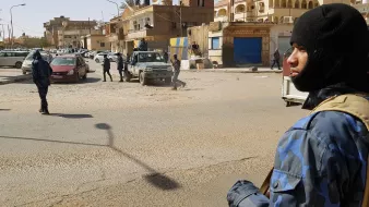 The image depicts a street scene in an urban environment where armed security personnel are present. One individual is prominently featured in the foreground, wearing a dark mask and tactical gear, holding a firearm. In the background, other security personnel are visible, some moving and others standing near vehicles, including a jeep. The street appears to be busy, with buildings lining either side and several parked cars. The setting suggests heightened security or law enforcement activity.