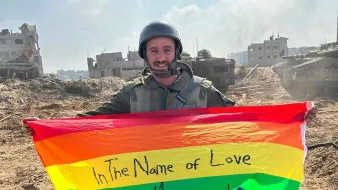 Un soldat souriant tient un drapeau arc-en-ciel sur un terrain dévasté.