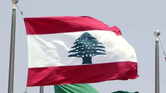 L'image montre deux drapeaux flottant côte à côte. Le drapeau du Liban est en haut, avec une bande rouge en haut et en bas, et une grande illustration d'un cèdre vert au centre. En dessous, le drapeau de l'Arabie Saoudite est vert, portant une inscription en arabe et une épée blanche. Les drapeaux sont hissés sur des mats, probablement lors d'un événement officiel ou d'une rencontre diplomatique.