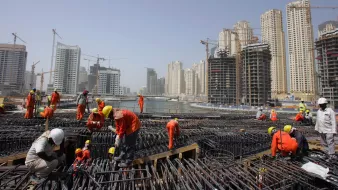 L'image montre un chantier de construction animé, où de nombreux ouvriers en uniformes orange et casques de sécurité s'affairent autour de structures en acier. En arrière-plan, on peut voir des immeubles modernes et des grues, illustrant une zone urbaine en développement. Le ciel est clair, et l'ensemble dégage une atmosphère dynamique de construction.