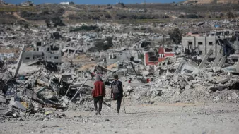 Due persone camminano tra le macerie di edifici distrutti in un paesaggio desolato.