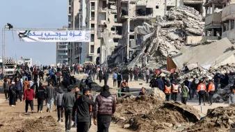 The image depicts a scene in Gaza featuring a street filled with people walking. In the background, there are buildings that appear to be damaged or destroyed. A large banner stretches across the street, welcoming visitors to Gaza, and some individuals wearing orange vests are visible, likely indicating they are part of a relief or aid organization. The area is marked by piles of debris and dirt, suggesting recent conflict or disaster. Overall, the atmosphere conveys a mix of resilience amid the challenges faced by the community.