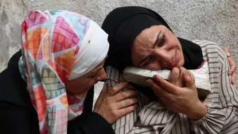 Two women in headscarves are embracing, expressing deep sorrow, with one holding a shoe.