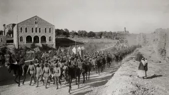 The image depicts a group of soldiers marching along a dirt pathway, likely in a historic setting. They are mounted on horses and are arranged in formation, indicating a military procession. In the background, there are stone buildings and a green landscape, suggesting a rural or semi-urban environment. Some individuals are also present on foot, possibly observing the parade. The overall tone of the photograph is sepia, contributing to its historical feel.