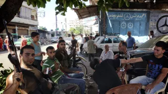 L'image montre un groupe d'hommes assis dans une rue animée. Certains d'entre eux portent des vêtements décontractés et semblent discuter ou se détendre ensemble. On peut voir un homme armé avec une arme à feu, ce qui suggère une ambiance tendue ou de conflit. En arrière-plan, il y a des voitures et des bâtiments, ainsi qu'une enseigne avec le logo d'une marque automobile. L'atmosphère semble informelle, malgré la présence d'armes.