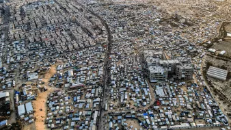 L'immagine mostra un'ampia area urbana vista dall'alto, caratterizzata da una fitta aggregazione di edifici e strutture. Si nota una forte densità abitativa, con molte abitazioni di dimensioni varie disposte in modo irregolare. Le strade principali e i passaggi sono visibili tra le varie costruzioni. In alcune zone si possono notare anche tende o strutture provvisorie, suggerendo la presenza di comunità temporanee. Sullo sfondo, si intravedono aree di terreni aperti e sviluppo urbano in espansione. La scena complessiva trasmette una sensazione di vivacità e organizzazione spontanea, con una tipologia abitativa che riflette una realtà complessa.