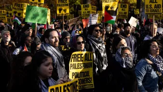 L'image montre une manifestation avec un grand nombre de personnes tenant des pancartes. Les pancartes portent des messages tels que "FREE PALESTINE" et "FREE ALL PALESTINIAN POLITICAL PRISONERS". Les participants sont vêtus de manière variée, certains portent des écharpes ou des keffiehs. On peut voir des drapeaux palestiniens en arrière-plan. L'ambiance semble être énergique et engagée, reflétant un fort sentiment de solidarité et de revendication pour les droits des Palestiniens.