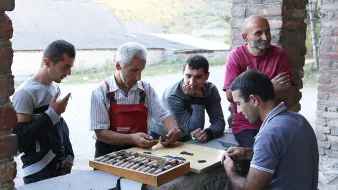 L'image montre un groupe de cinq hommes rassemblés autour d'une table en pierre en extérieur. Sur la table, il y a un plateau avec des pièces de jeu, et un homme au centre semble expliquer ou démontrer quelque chose. Les autres hommes écoutent attentivement, certains les bras croisés et d'autres penchés vers la table. Le décor naturel en arrière-plan présente des collines verdoyantes, ce qui suggère un environnement rural. L'atmosphère semble conviviale et détendue.