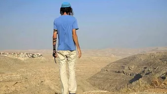 Cette image montre une personne de dos, se tenant au bord d'une falaise. Elle porte un t-shirt bleu et des pantalons clairs, ainsi qu'une kippa sur la tête. Son bras gauche est vêtu d'un bandage. Le paysage environnant est désertique, avec des collines et une vallée visible à l'arrière-plan. On peut apercevoir quelques maisons au loin, suggérant une présence humaine dans cette vaste étendue. L'atmosphère est calme, avec un ciel clair.