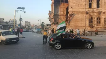 Scène de rue avec un bâtiment endommagé, des gens et une voiture avec un drapeau.