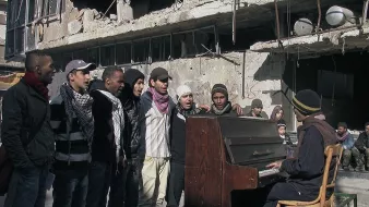 L'image montre un groupe de jeunes hommes chantant autour d'un piano dans un environnement dégradé, probablement après un conflit. Les bâtiments en arrière-plan sont en ruines, témoignant de destructions significatives. Les chanteurs semblent engagés et déterminés, tandis qu'un homme joue du piano, créant une atmosphère de solidarité et d'espoir au milieu de la désolation.