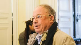 Homme âgé avec des lunettes, portant un manteau, dans un intérieur lumineux.