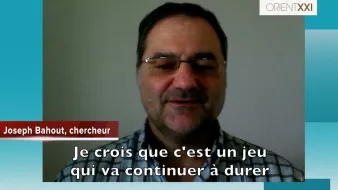 L'image montre un homme apparaissant en vidéoconférence. Il porte des lunettes et un chemisier à carreaux. Le texte en bas de l'image indique son nom, "Joseph Bahout, chercheur", suivi d'une citation où il exprime son opinion sur une situation, affirmant qu'il croit que "c'est un jeu qui va continuer à durer". Le fond est simple, et l'interface semble être d'une plateforme de communication vidéo.