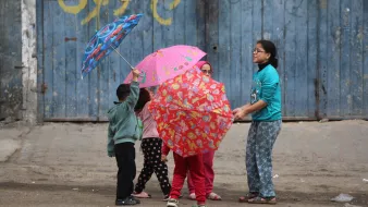 L'image montre un groupe d'enfants jouant dans la rue. Ils ont des parapluies colorés, certains avec des motifs amusants. Les enfants semblent joyeux et engagés dans leur jeu. En arrière-plan, il y a un mur bleu, qui donne une ambiance urbaine à la scène. Ils portent des vêtements confortables, adaptés à une activité en extérieur. L'atmosphère générale est légère et pleine de vie.