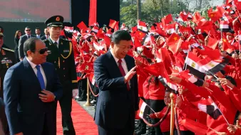 L'image montre un événement public avec des drapeaux de la Chine et de l'Égypte. Au premier plan, deux figures politiques semblent interagir avec la foule. L'un des hommes porte un costume sombre et des lunettes de soleil, tandis que l'autre est habillé d'un costume clair et sourit en saluant les personnes présentes. La foule, vêtue de vêtements rouges, tient des drapeaux et semble enthousiaste. On peut voir un tapis rouge et des gardes en uniforme à l'arrière-plan. L'ambiance est festive et accueillante.