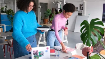 En la imagen se pueden ver a dos mujeres en un espacio de trabajo moderno y luminoso. Una de ellas, con cabello rizado y vestido azul, está sosteniendo unos cuadernos o carpetas, mientras que la otra, con cabello rizado y recogido, está organizando unos libros sobre una mesa. En la mesa hay una computadora portátil, algunos materiales impresos y una planta decorativa. El ambiente parece creativo y acogedor, con elementos de diseño contemporáneo y luminosidad natural.