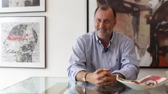 L'image montre un homme assis à une table en verre, semblant détendu et souriant. Il porte une chemise à rayures et des bracelets. En arrière-plan, on peut voir des œuvres d'art accrochées au mur, donnant une ambiance artistique à la pièce. Sur la table, il y a une assiette et des documents, ce qui suggère un contexte de discussion ou d'entretien. L'ensemble donne une impression de convivialité et d'élégance.